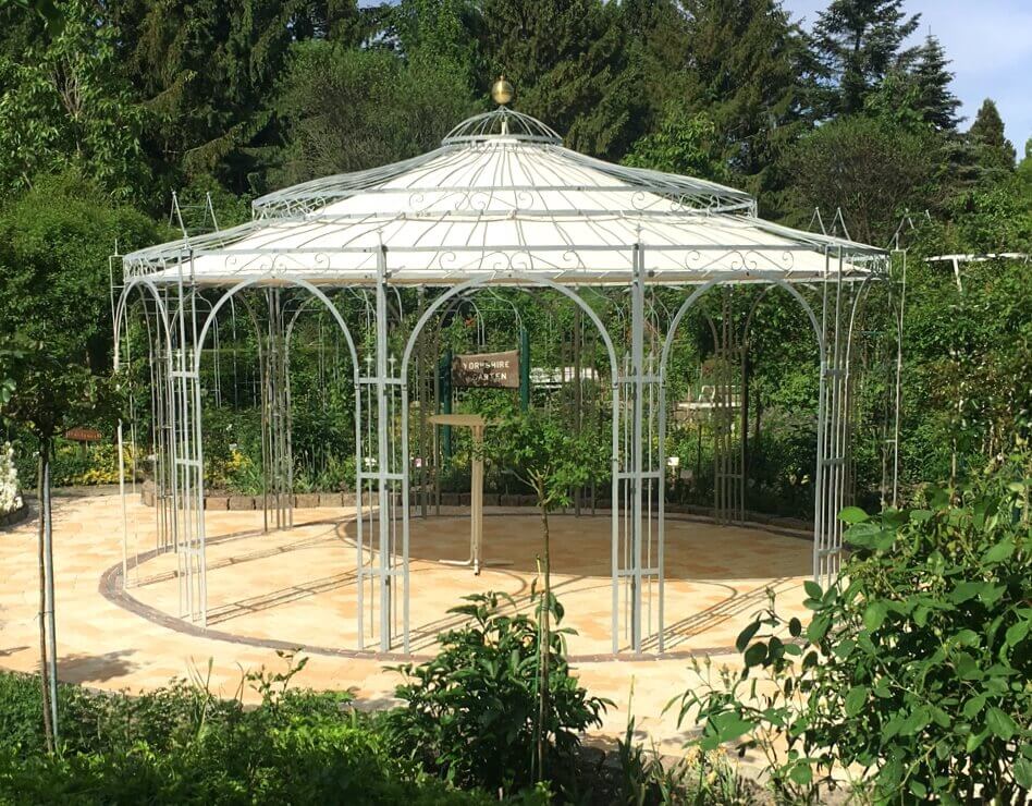 Pavillon Toskana in feuerverzinkter Ausführung mit Sonnensegel und Messingkugel in einem bewachsenen Showgarten