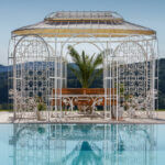 Pavillon Verona in pulverbeschichteter Sonderfarbe mit Sonnensegel und Rankgitter hinter modernen Pool