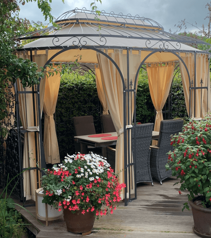 Pavillon Verona in pulverbeschichteter Ausführung mit Sonnensegel und Vorhängen auf einer Holzerhöhung mit Essenstisch und Stühlen