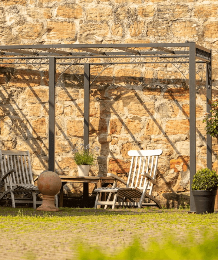Pergola Sardegna in pulverbeschichteter Ausführung mit Sonnensegel an Sandsteinmauer mit Holzliegen
